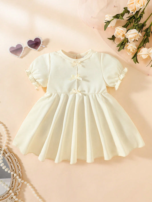 Prenda beige vestido para bebe con lazos decorativos vista frontal
