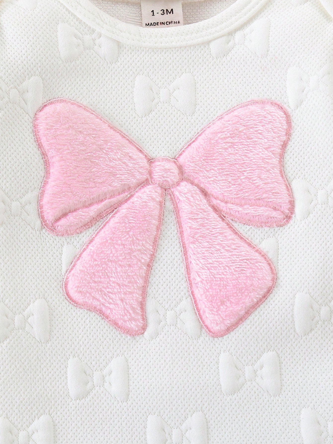 Prenda blanco mameluco para bebe detalle de lazo rosado en relieve

