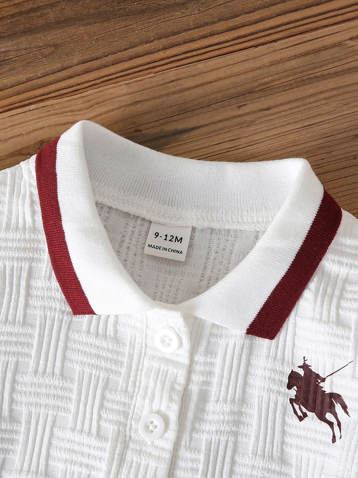 Camisa polo blanca para bebé con cuello burdeos, botones frontales y textura geométrica.