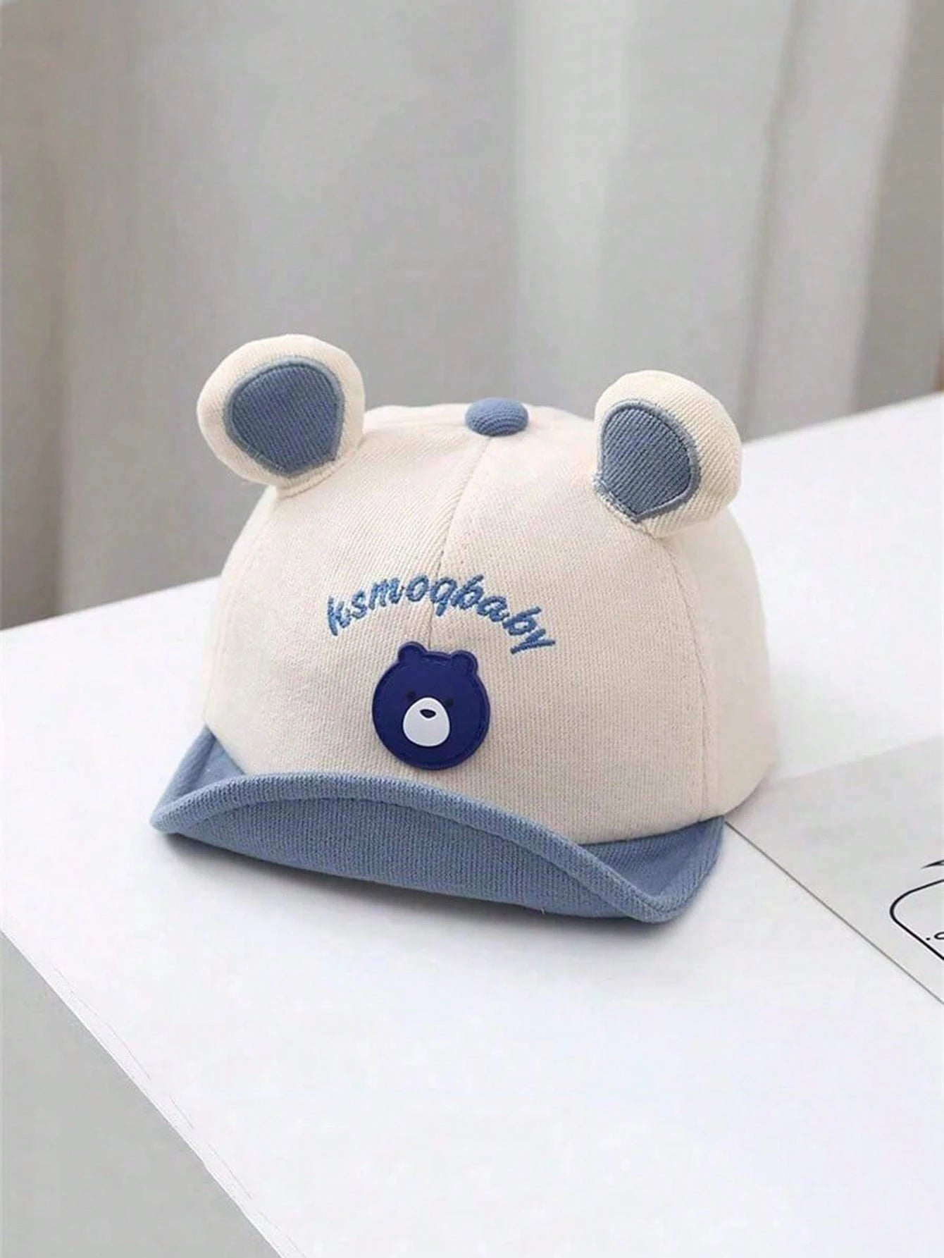 Gorro beige para bebe con orejitas azules y visera azul vista frontal
